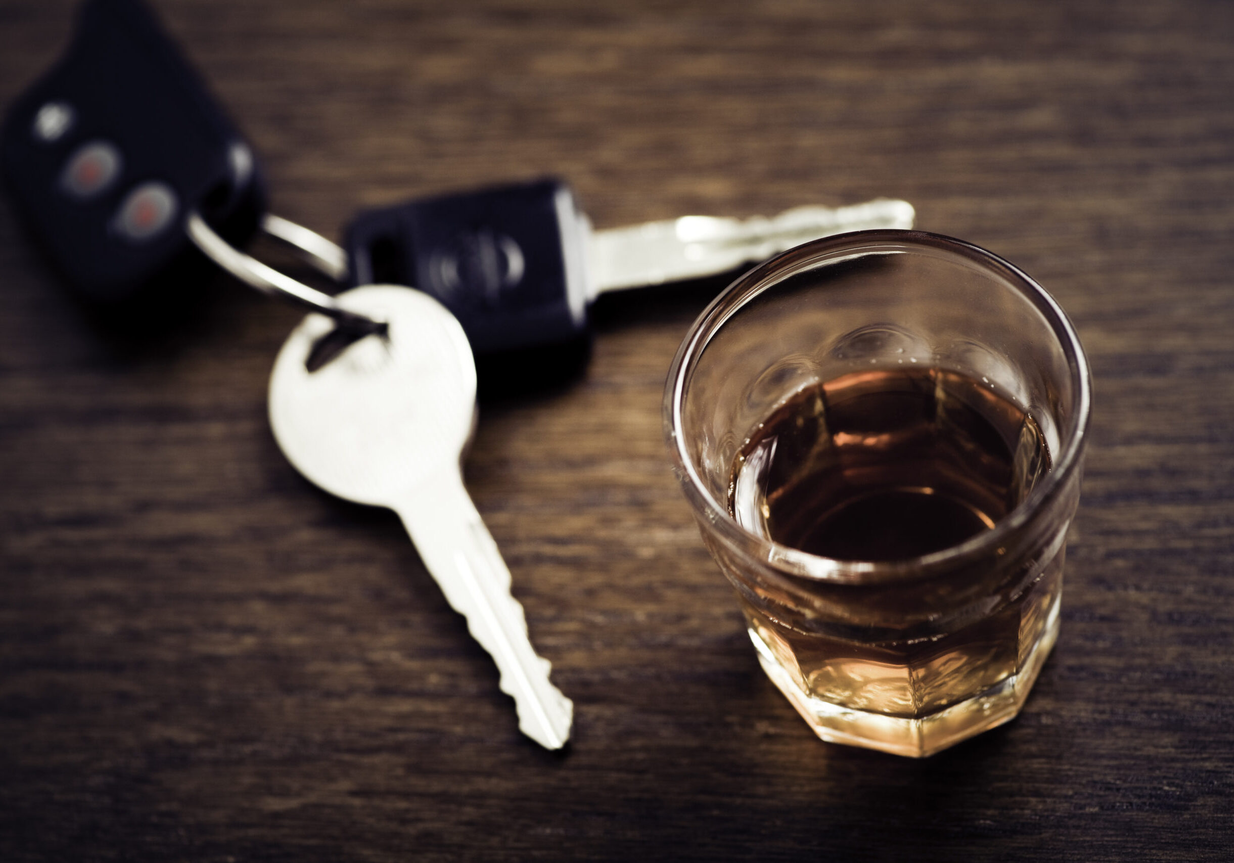 A glass of whisky and a set of car keys on a wooden table A shot glass full of dark colored alcohol on top of a bar table along with a set of car keys. Drinking and driving series. Also driving under the influence of alcohol.